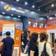 Trade show booth for AP Systems with orange displays and attendees walking by; large signage overhead reads AP Systems and stock code 688348, booth 1-E21.