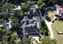 Aerial view of a large gray-roofed house with a glass-enclosed back area, surrounded by trees and a curved driveway.
