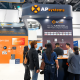 Busy AP Systems trade show booth with curved blue signage, orange logo, and attendees gathered around a counter for product demos and information marketing materials in a large expo hall.